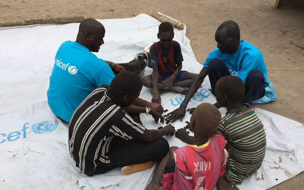 Durch das Spielen werden die Kinder wieder zusammengebracht und bilden Gemeinschaften | © UNICEF/Michael Charley Südsudan: Michael sitzt mit einem Ball auf einer Plane und spricht mit Kindern