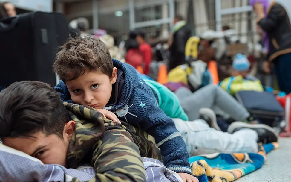 Migration: Millionen von Kindern und Jugendlichen sind weltweit Schutzlosigkeit und Gewalt ausgeliefert. | UNICEF/Arcos Ein Kind liegt auf dem Rücken seines großen Bruders in einem Saal wartender Migranten