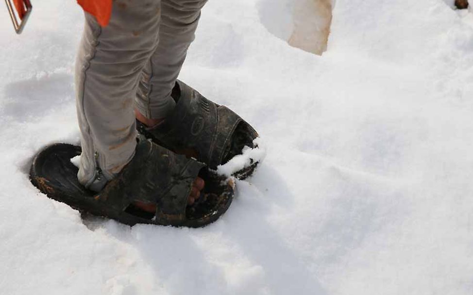 Ein Flüchtlingskind steht in viel zu großen Sandalen im Schnee.