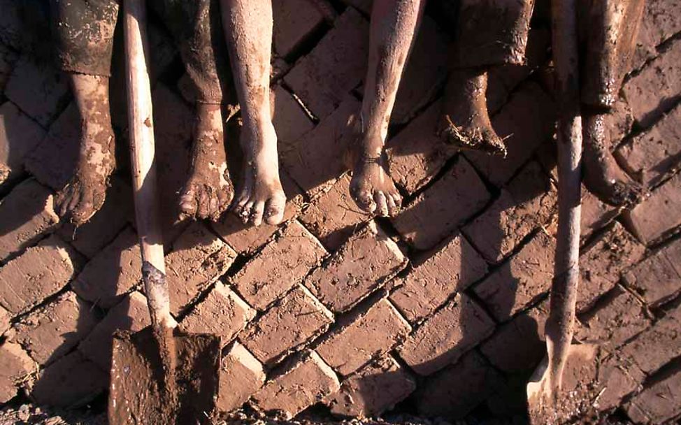 Irak: Die schlammigen Füße von drei Kindern hängen von einer Backsteinmauer, an der zwei Spaten lehnen.