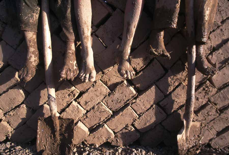 Irak: Die schlammigen Füße von drei Kindern hängen von einer Backsteinmauer, an der zwei Spaten lehnen.