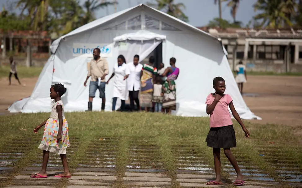 UNICEF richtet Zelte für die medizinische Versorgung in Buzi/Mosambik ein. | © UNICEF/Prinsloo Zwei Kinder im Vordergrund laufen an einem UNICEF-Zelt im Hintergrund vorbei, aus dem zwei Ärzte kommen.