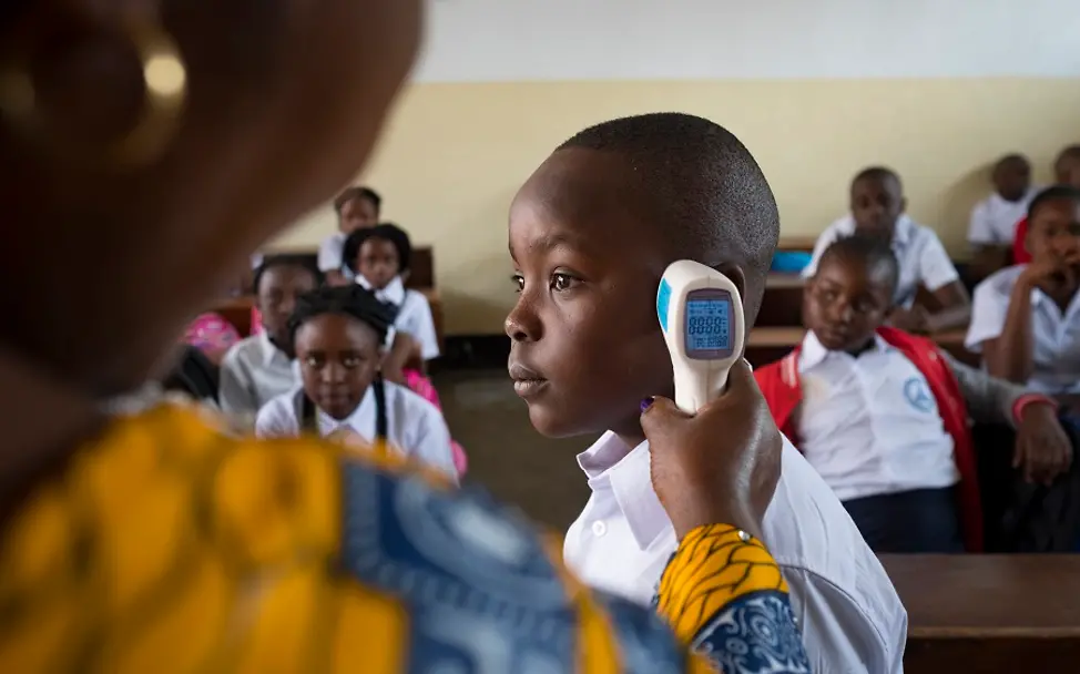 Mit einem digitalen Thermometer kann die Temperatur der Schülerinnen und Schüler überprüft werden. | © UNICEF/Nybo Eine Lehrerin hält einem Schüler vor der Klasse ein digitales Thermometer an den Kopf.