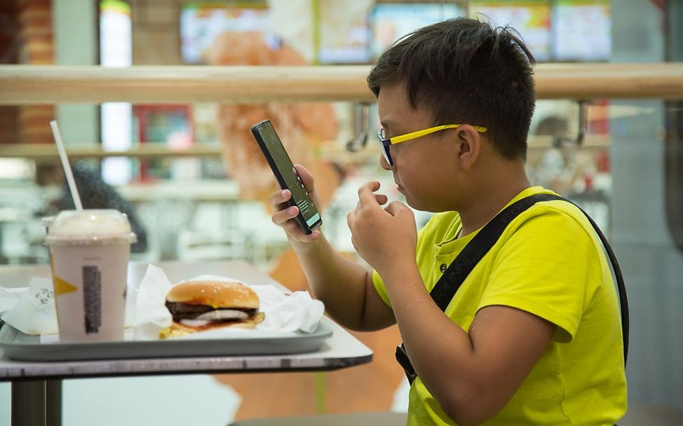 Ein Junge schaut auf sein Smartphone während er sein Fast Food Menü isst.