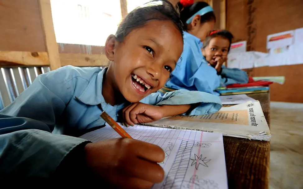 Manjari Tamang (7) aus Nepal. | © UNICEF/Karki Nepal: Ein lachendes Mädchen sitzt an einem Pult und schreibt in ein Heft.