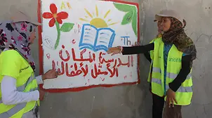 Muzoon vor einem Bild, das Kindern zeigen soll, wie wichtig Schulbildung ist. | © UNICEF/Fricker Jordanien: Muzoon und zwei weitere UNICEF-Helfer betrachten ein Bild zum Thema Schulbildung.