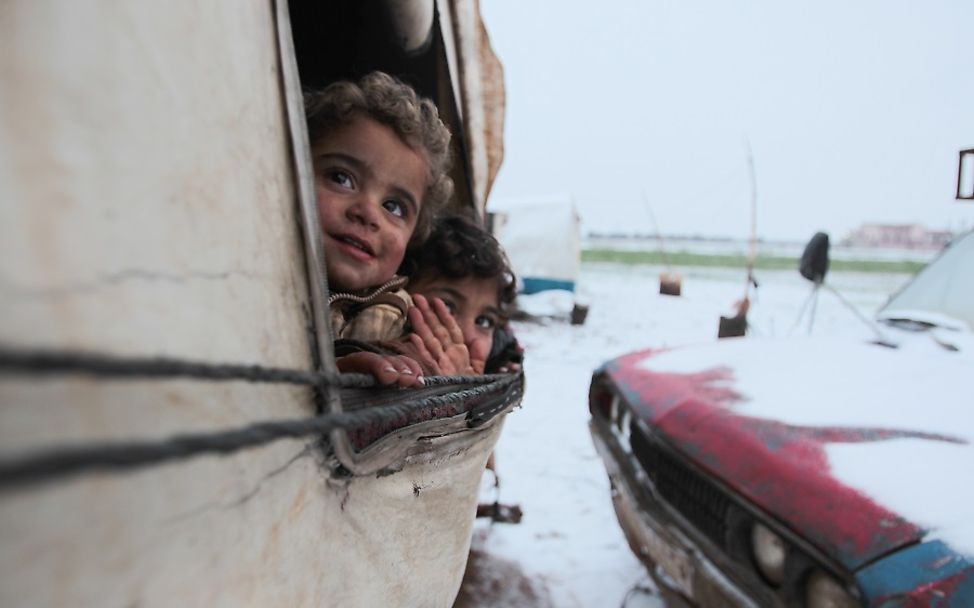 Zwei Kinder schauen aus einem Zelt auf ein verschneites Flüchtlingslager.