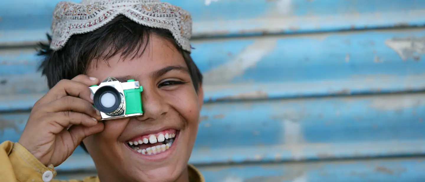 Ein Junge aus Pakistan hält eine kleine Spielzeug-Kamera vor sein Auge und lacht.