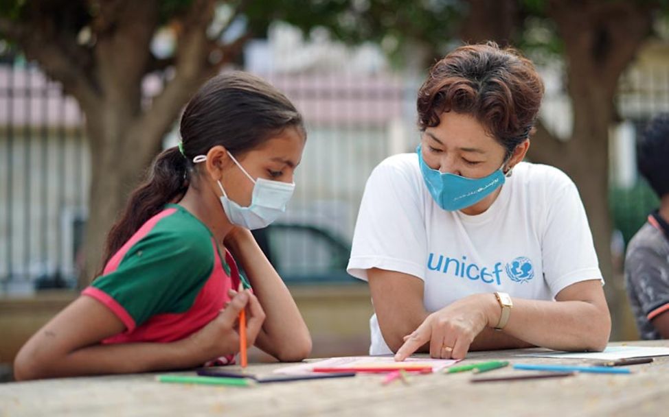 Ein Mädchen im Libanon sitzt mit einer UNICEF-Helferin draußen an einem Arbeits- und Maltisch.