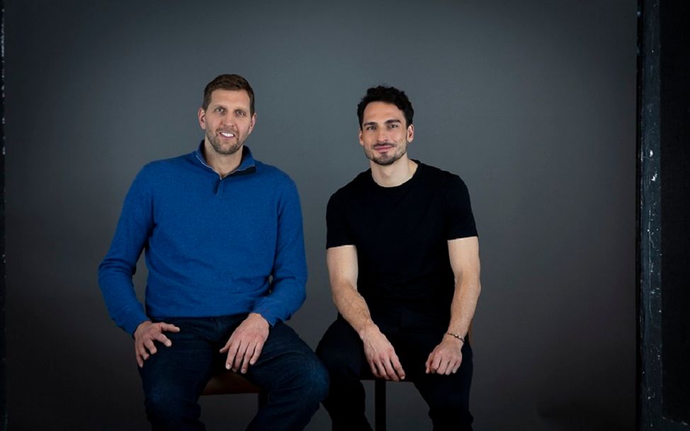 Dirk Nowitzki und Mats Hummels sitzen auf zwei Stühlen und schauen in die Kamera.