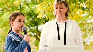 Kinderrechteschulen: Eine Schülerin mit einem Mikrofon in der Hand steht mit der Schulleiterin vor einem Pult.