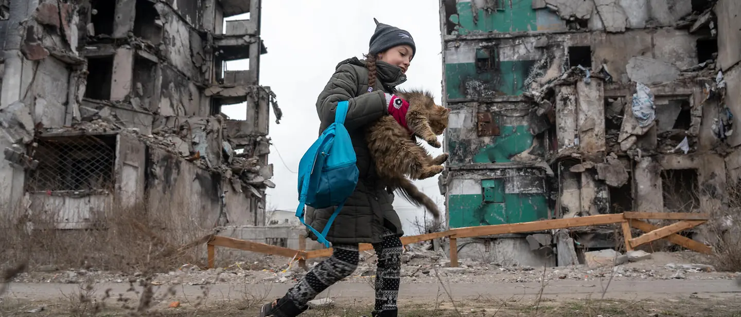 Ein junges Mädchen mit einer Katze im Arm vor ihrem ehemaligen, nun zerbombten Zuhause