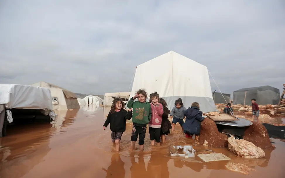 Kinder stehen in einem überfluteten Gebiet des Lagers Kafr Losin im Nordwesten von Syrien | © UNICEF/UN0405689/Akacha Weltflüchtlingstag: Kinder stehen in einem überfluteten Gebiet des Lagers Kafr Losin im Nordwesten von Syrien.