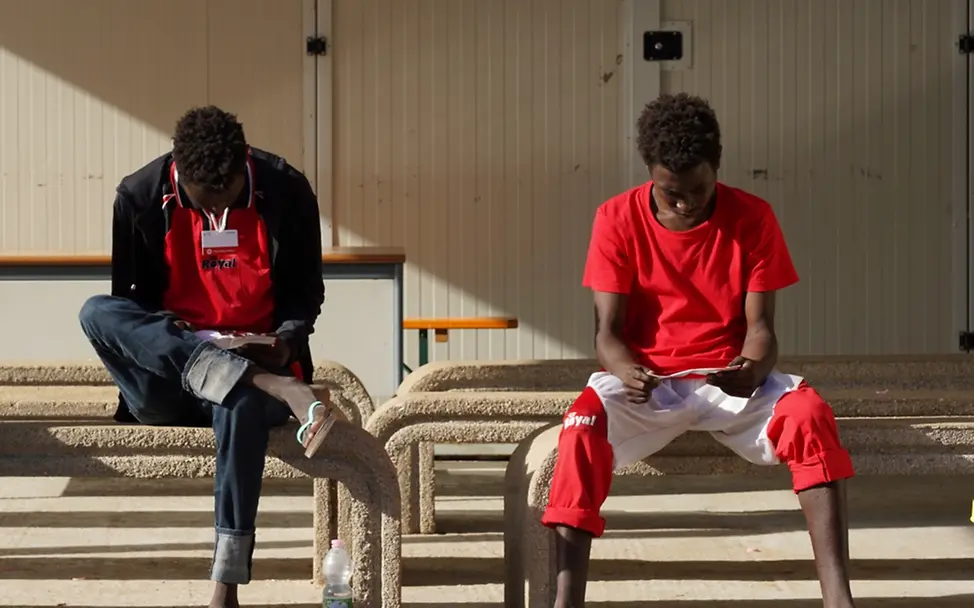 Unbegleitete jugendliche Migranten sitzen im "Hotspot"-Aufnahmezentrum auf der Insel Lampedusa, Italien, zusammen und lesen Informationsbroschüren | © UNICEF/UNI443174/Corti Weltflüchtlingstag: Jugendliche Migranten lesen im Aufnahmezentrum der Insel Lampedusa, Italien, Informationsbroschüren.