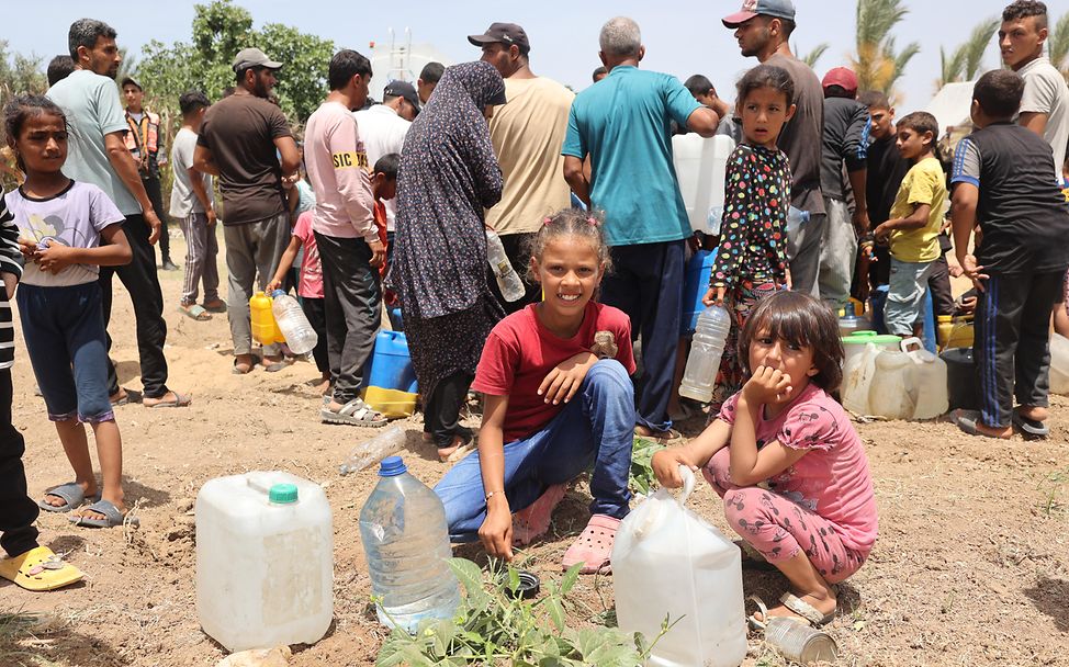 Zwei Mädchen hocken auf dem Boden, vor ihnen gefüllte Wasserkanister, hinter ihnen viele Menschen ebenfalls mit Kanistern