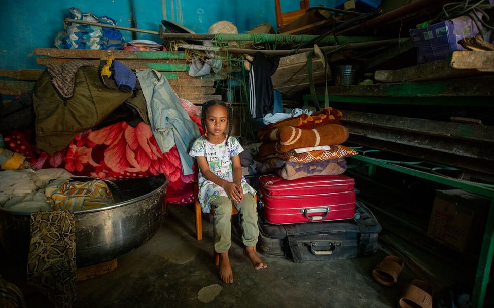 A little girl sits on a chair surrounded by suitcases and utensils in her new home