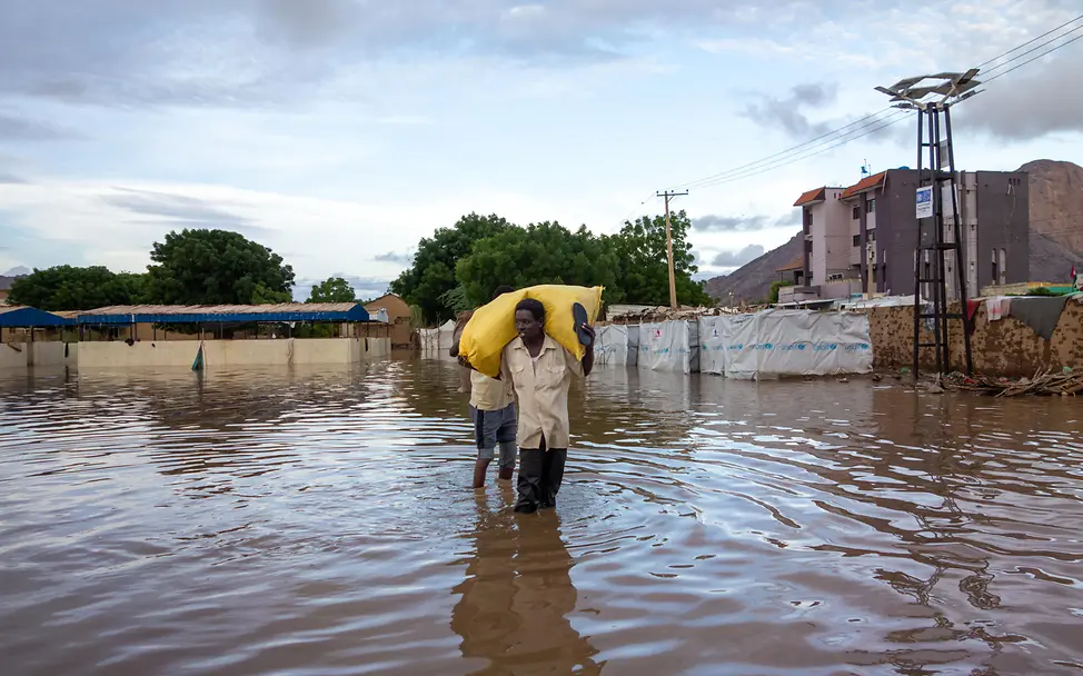 Fluten im Sudan erschweren das Leben zusätzlich | © UNICEF/UNI618360/Alfadil Eine Person mit einem Sack auf den Schultern geht durch kniehohes Wasser