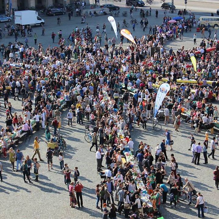 Auf einem großen Platz sind sternförmig lange Tafeln aufgebaut, viele Menschen stehen dabei 