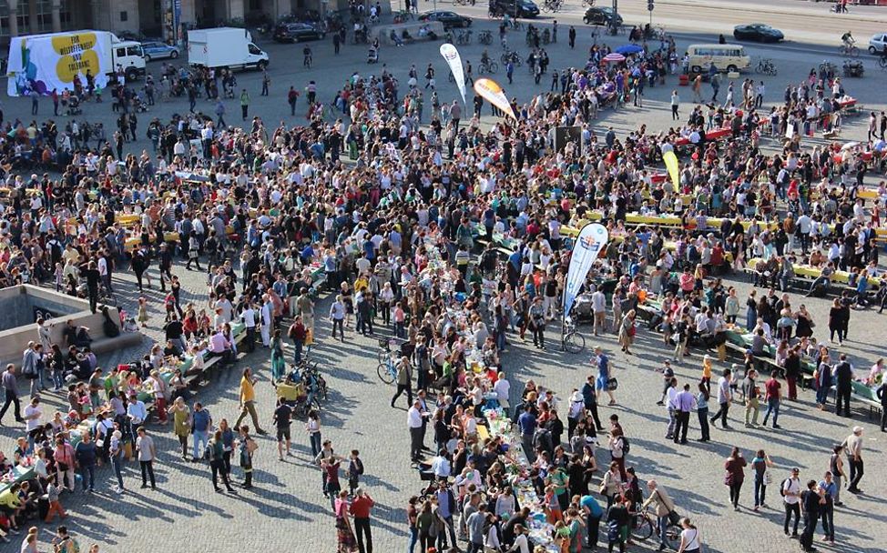 Auf einem großen Platz sind sternförmig lange Tafeln aufgebaut, viele Menschen stehen dabei 