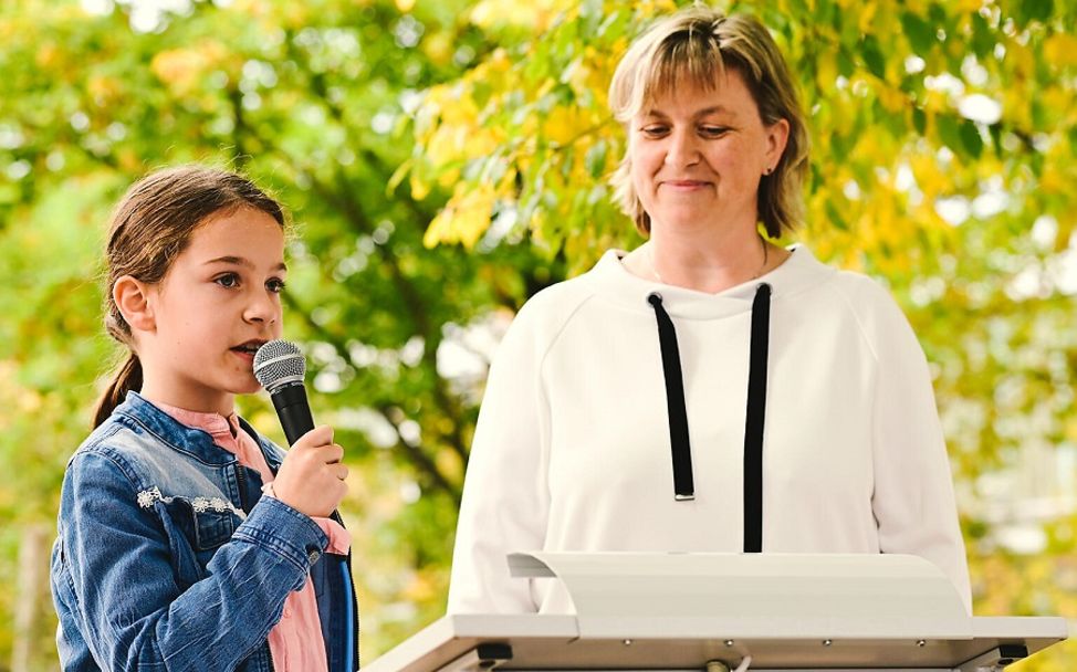 Kinderrechteschulen: Eine Schülerin mit einem Mikrofon in der Hand steht mit der Schulleiterin vor einem Pult.