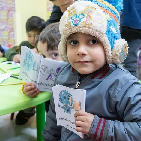 Der kleine Ghaith in einem Kinderschutzzentrum von UNICEF | UNICEF/UNI704243/Yacoubian Ein kleines Kind hält ein gemaltes Bild in die Kamera