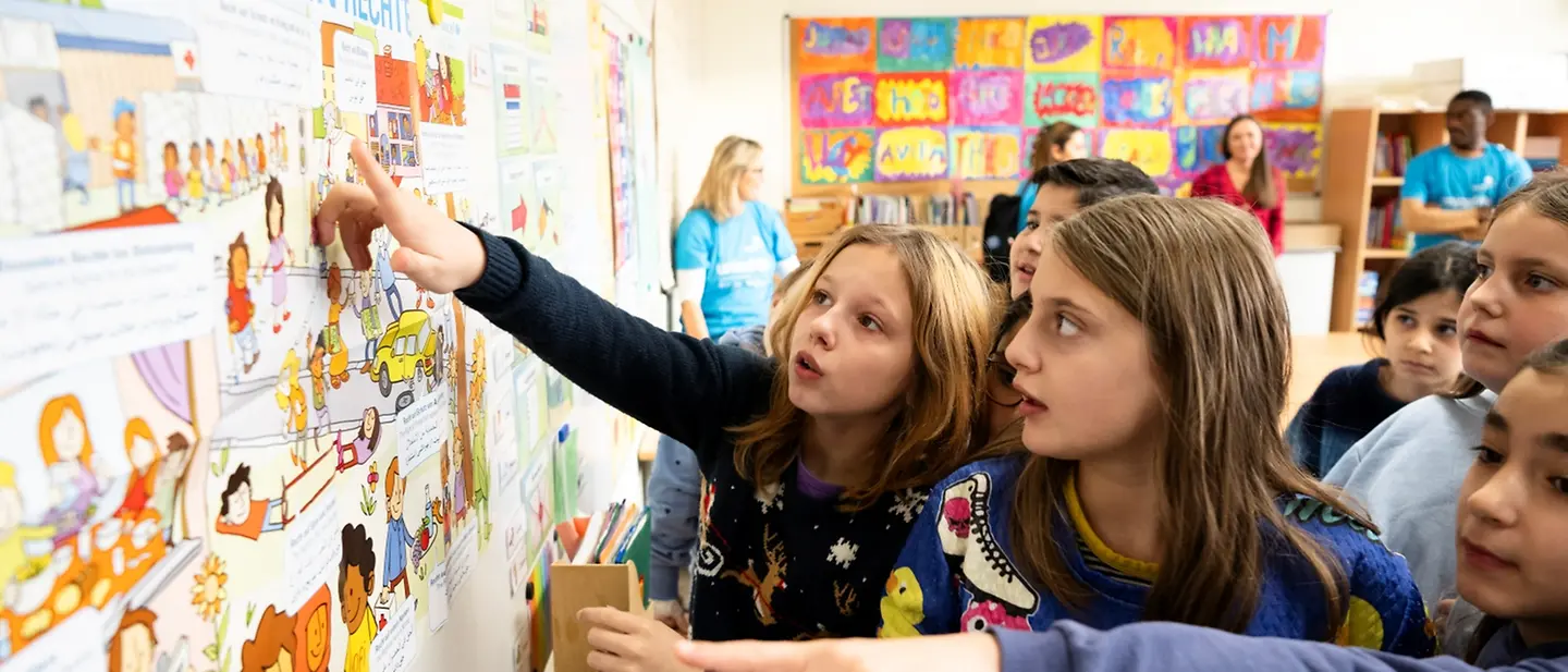 Kinder einer Schulklasse stehen vor dem Plakat mit den Rechten des Kindes und zeigen auf das Plakat.