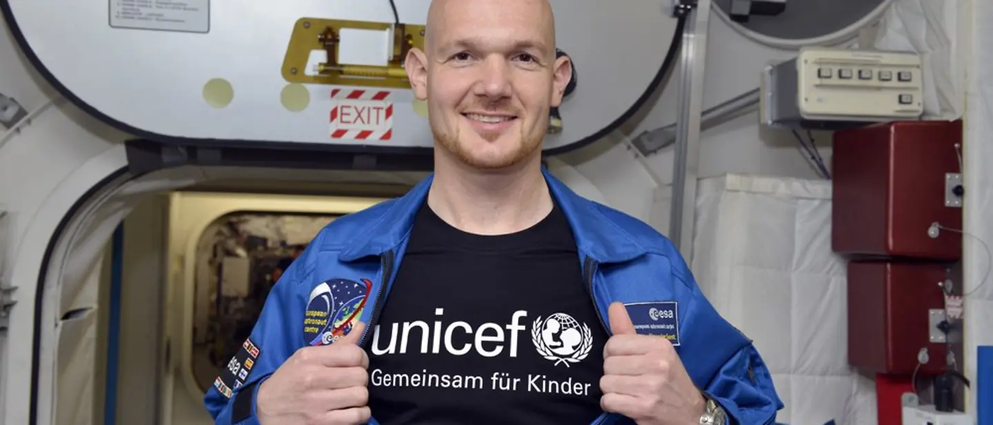 Der Astronaut Alexander Gerst steht in einem Raumschiff und öffnet seine Jacke, worunter ein UNICEF T-Shirt zum Vorschein kommt.